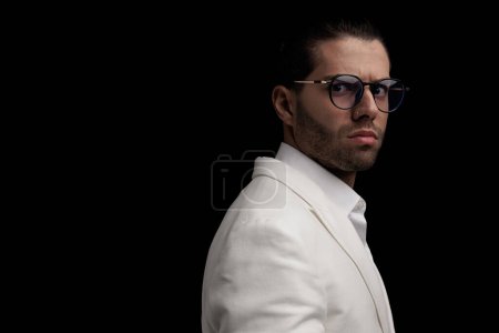 Téléchargez les photos : Vue latérale de l'homme élégant cool avec des lunettes portant un costume blanc et posant devant un fond noir - en image libre de droit