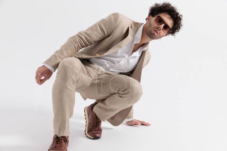 Téléchargez les photos : Sexy smart casual guy portant des lunettes de soleil accroupi et toucher le sol en face de fond gris en studio - en image libre de droit