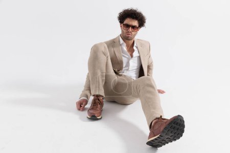 Photo for Handsome guy with curly hair sitting on the floor with on knee up and posing in a cool way on grey background - Royalty Free Image