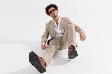 Téléchargez les photos : Attrayant homme élégant avec chemise col ouvert et costume beige assis sur le sol sur fond gris - en image libre de droit