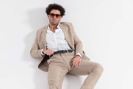Téléchargez les photos : Homme de mode sexy avec chemise col ouvert couché sur le sol et posant en toute confiance tout en ajustant la ceinture sur fond gris - en image libre de droit