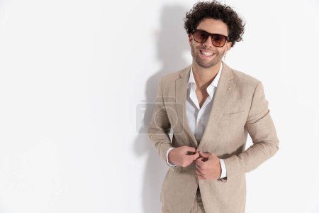 Téléchargez les photos : Portrait d'homme décontracté intelligent heureux avec chemise col ouvert riant et déboutonnant costume beige devant fond gris en studio - en image libre de droit