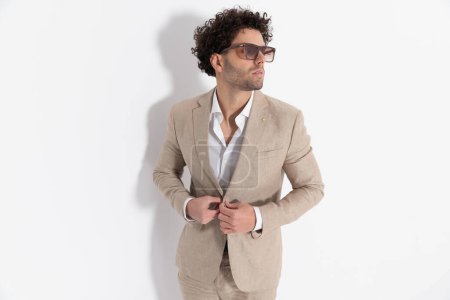 Téléchargez les photos : Homme décontracté intelligent avec chemise col ouvert regardant vers le côté et boutonnage costume beige devant fond gris en studio - en image libre de droit