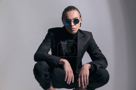Téléchargez les photos : Photo de mode de jeune homme d'affaires accroupi et posant avec une ambiance cool et portant une belle tenue sur fond de studio gris - en image libre de droit