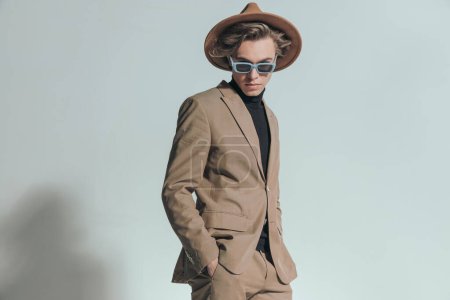 Téléchargez les photos : Jeune homme de mode avec des lunettes de soleil et chapeau posant avec les mains dans les poches en face de fond gris - en image libre de droit