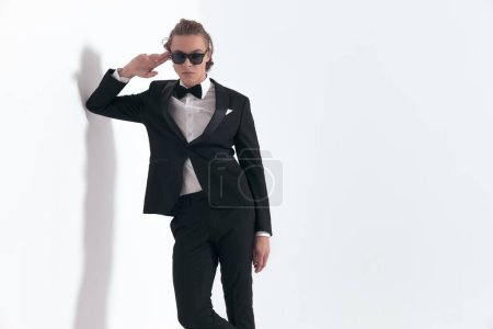 Téléchargez les photos : Homme d'affaires élégant confiant aux cheveux longs et smoking posant son coude sur le mur, ajustant les lunettes de soleil et posant sur fond gris - en image libre de droit