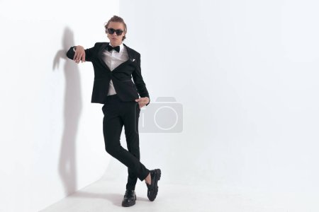 Téléchargez les photos : Marié élégant avec des lunettes de soleil tenant son coude sur le mur et posant avec la main dans les poches d'une manière confiante en face de fond gris - en image libre de droit