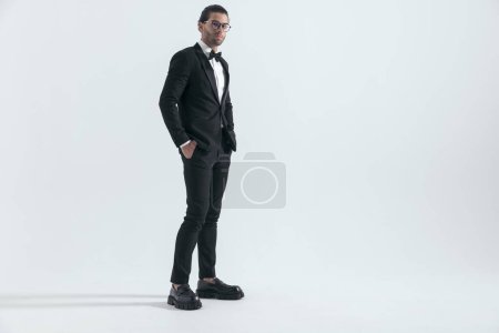 Photo for Side view of elegant man in tuxedo holding hands in pockets and smiling in front of grey background - Royalty Free Image