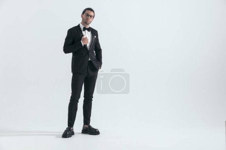 Photo for Sexy young man with glasses adjusting black tuxedo and posing with hand in pockets while standing on grey background - Royalty Free Image
