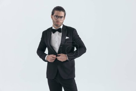 Photo for Attractive elegant man with glasses closing and buttoning black tuxedo in front of grey background in studio - Royalty Free Image
