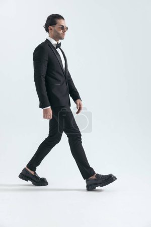 Photo for Cool elegant man with sunglasses in black tuxedo looking to side and walking in front of grey background in studio - Royalty Free Image