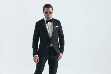 Photo for Sexy young guy in black tuxedo wearing sunglasses and frowning in front of grey background - Royalty Free Image