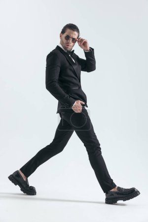 Foto de Novio de moda fresco en esmoquin negro ajustando gafas de sol y caminando con la mano en el bolsillo delante de fondo gris en el estudio - Imagen libre de derechos