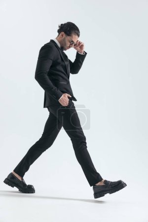 Photo for Side view of cool elegant man in tuxedo walking with hand in pocket, looking down and adjusting sunglasses in front of grey background in studio - Royalty Free Image