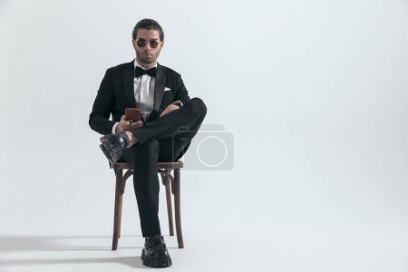 Téléchargez les photos : Homme d'affaires confiant en smoking noir tenant mobile et textos assis sur fond gris - en image libre de droit