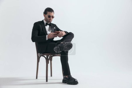 Téléchargez les photos : Attrayant homme élégant avec des lunettes de soleil défilant sur les médias sociaux tout en étant assis sur fond gris - en image libre de droit
