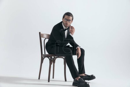 Téléchargez les photos : Élégant jeune homme d'affaires avec des lunettes assis avec des coudes sur les cuisses et posant devant fond gris - en image libre de droit