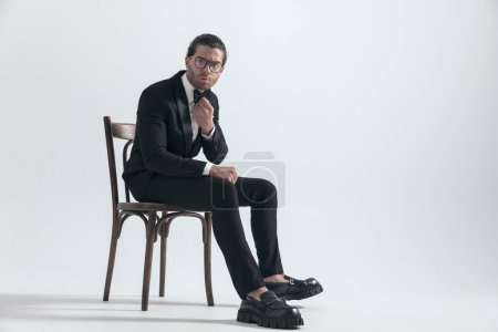Téléchargez les photos : Homme élégant sérieux en smoking noir fronçant les sourcils et fixant noeud papillon tout en étant assis sur une chaise en bois sur fond gris - en image libre de droit