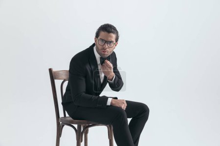 Téléchargez les photos : Vue latérale de cool homme élégant avec des lunettes assis et réglage noeud papillon devant fond gris - en image libre de droit