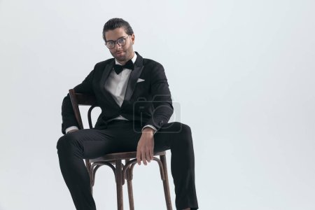 Téléchargez les photos : Heureux jeune homme élégant avec des lunettes portant smoking noir assis sur une chaise en bois et souriant sur fond gris - en image libre de droit