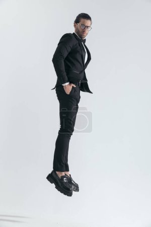Photo for Elegant groom in black tuxedo jumping in the air with hands in pockets in front of grey background - Royalty Free Image