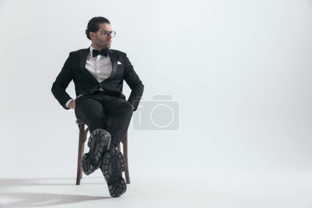 Téléchargez les photos : Sexy jeune homme en smoking noir avec des lunettes assis avec les mains dans les poches et regardant de côté sur fond gris - en image libre de droit