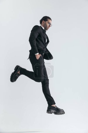 Photo for Side view of cool fashion guy looking to side and jumping with hands in pockets in front of grey background - Royalty Free Image