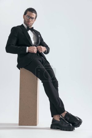 Téléchargez les photos : Sexy jeune marié avec des lunettes assis et déboutonner smoking noir en face de fond gris - en image libre de droit