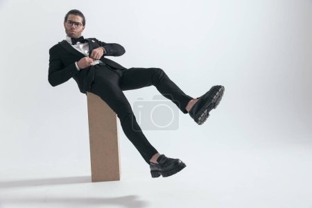 Téléchargez les photos : Jeune marié sexy avec des lunettes boutonnage costume tout en étant assis avec les jambes en l'air sur fond gris en studio - en image libre de droit