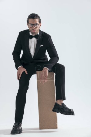 Foto de Sexy hombre de moda con gafas en esmoquin negro posando con los brazos en los muslos sobre fondo gris - Imagen libre de derechos
