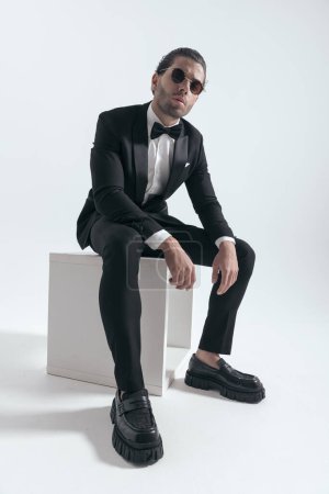 Téléchargez les photos : Confiant homme élégant avec des lunettes de soleil assis avec les bras sur les genoux et posant d'une manière fraîche sur fond gris - en image libre de droit