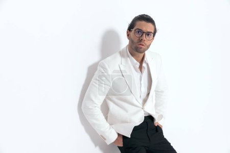 Téléchargez les photos : Confiant homme sexy tenant la main dans les poches et reposant sur un mur en face de fond gris en studio - en image libre de droit