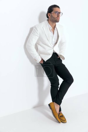 Foto de Hombre sexy fresco con gafas con traje de chaqueta blanca, tomados de la mano en los bolsillos y acostados en una pared frente al fondo gris en el estudio - Imagen libre de derechos