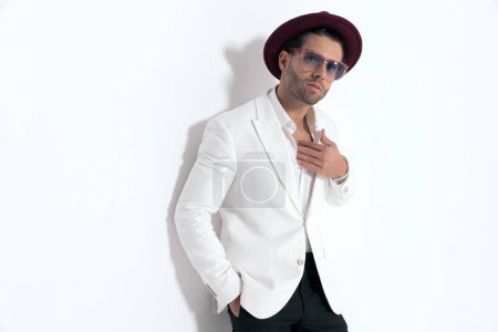 Téléchargez les photos : Sexy fashion guy avec chapeau et lunettes de soleil tenant la main dans les poches, ajustant chemise à col ouvert et couché sur un mur sur fond gris - en image libre de droit