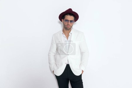 Foto de Hombre elegante guapo con sombrero y gafas, con traje de chaqueta blanca y camisa de cuello abierto, cogidas de la mano en bolsillos sobre fondo gris - Imagen libre de derechos