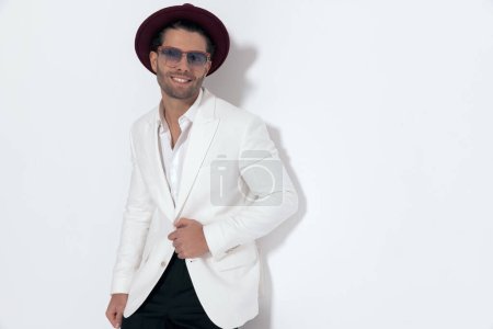 Téléchargez les photos : Attrayant jeune homme avec chapeau et lunettes de soleil ajuster costume veste blanche et rire tout en étant allongé sur un mur sur fond gris - en image libre de droit