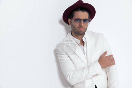 Téléchargez les photos : Cool jeune homme portant chapeau et lunettes de soleil tenant bras de manière mode et posant devant fond gris - en image libre de droit
