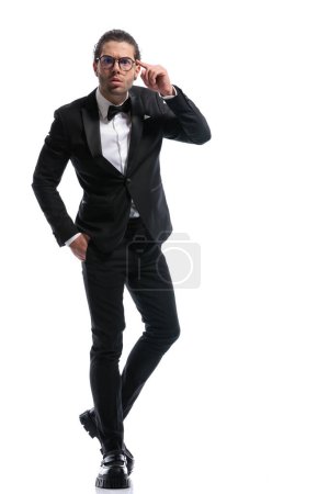 Téléchargez les photos : Attrayant homme d'affaires pointant la tête avec la main dans la poche, jambes croisées et portant des lunettes sur fond de studio blanc - en image libre de droit