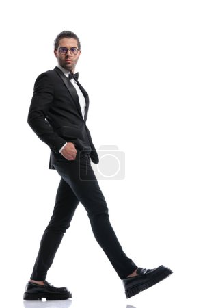 Téléchargez les photos : Attrayant homme d'affaires marchant de côté avec dans les poches et portant des lunettes sur fond de studio blanc - en image libre de droit