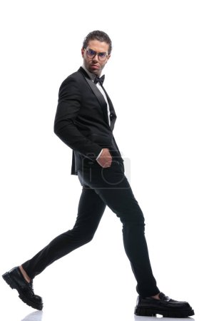 Téléchargez les photos : Jeune homme d'affaires marchant large avec les mains dans les poches et le visage fou et portant des lunettes sur fond de studio blanc - en image libre de droit