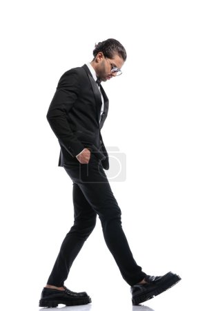 Téléchargez les photos : Bel homme d'affaires marchant les mains dans les poches et regardant vers le bas et portant des lunettes sur fond de studio blanc - en image libre de droit