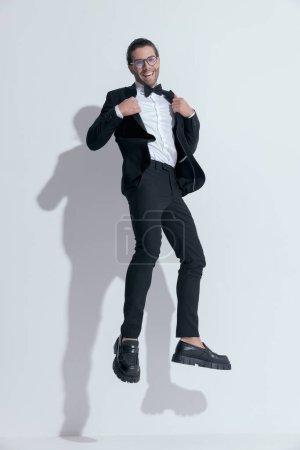 Foto de Atractivo hombre de negocios atrapado arreglando su esmoquin y riendo en el aire, usando gafas contra fondo blanco estudio - Imagen libre de derechos