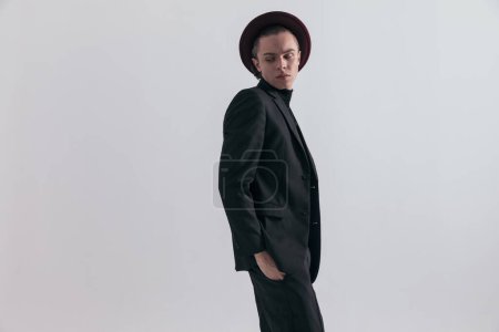 Foto de Imagen de moda de hombre de negocios sexy mirando hacia atrás con las manos en los bolsillos y usando un traje elegante contra el fondo gris del estudio - Imagen libre de derechos