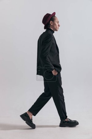 Foto de Imagen de moda de un joven hombre de negocios caminando con actitud dura y usando un atuendo elegante contra el fondo gris del estudio - Imagen libre de derechos