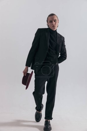 Foto de Imagen de moda de un joven hombre de negocios caminando hacia la cámara mientras se quita el sombrero y usa un atuendo elegante contra el fondo gris del estudio. - Imagen libre de derechos