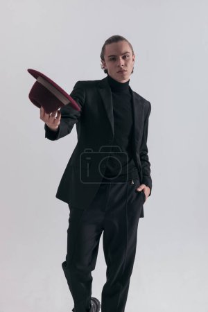 Foto de Imagen de moda de hombre de negocios sexy mostrando su sombrero en la cámara y usando un traje elegante contra fondo gris estudio - Imagen libre de derechos