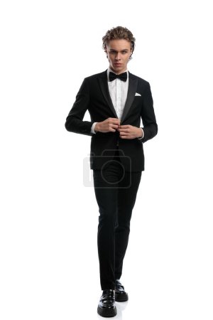 Foto de Sexy hombre de negocios caminando y cerrando su esmoquin, vistiendo un traje formal contra fondo de estudio blanco - Imagen libre de derechos