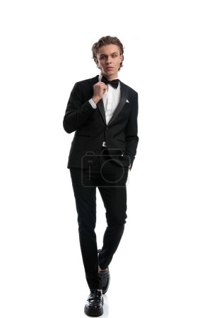Photo for Young businessman fixing bowtie with one hand in pocket, wearing a formal outfit against white studio background - Royalty Free Image