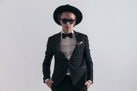 Photo for Portrait of elegant young groom with hat and sunglasses wearing black tuxedo and holding hands in pockets while posing in front of grey background in studio - Royalty Free Image