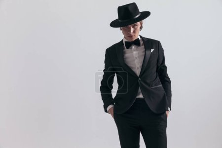 Téléchargez les photos : Élégant homme de mode en smoking noir avec chapeau tenant la main dans les poches devant fond gris en studio - en image libre de droit